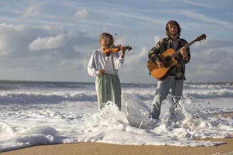Eine Frau mit Geige und ein ein Mann mit Gitarre stehen in der schäumenden Gischt am Meeresstrand