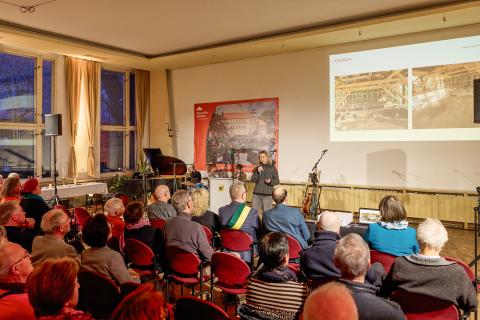 Publikum sitz bei einer Veranstaltung in Reihen und hört einer Sprecherin zu